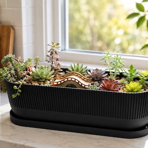 May include: A long, black, ribbed planter filled with various green and reddish succulents and a small, beige, arched bridge ornament. The planter sits on a matching black tray, containing light-coloured pebbles around the plants.
