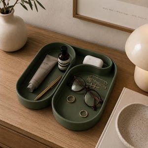 May include: A matte olive green ceramic tray with an organic, curved, multi-compartment shape. The tray holds a tube of cream, a small dark bottle, a gold pen, a white earbud case, a pair of dark-rimmed glasses, two gold rings, and a gold chain.
