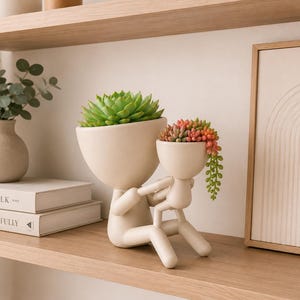 May include: Two cream ceramic planters in the shape of figures, one holding the other. The larger planter contains a green succulent, and the smaller one has a trailing succulent with red, orange, and green colours. The planters are on a wooden shelf.