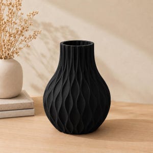May include: A matte black vase with a rounded, bulbous body and a narrow neck. The exterior surface features a repeating vertical wave pattern that creates deep, textured ridges around the entire circumference of the vessel.