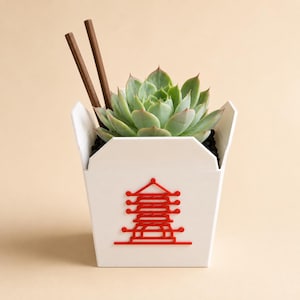 May include: A white takeout container-shaped planter featuring a red pagoda graphic on the front. The planter holds a green succulent plant and includes two brown wooden chopsticks protruding from the top, set against a solid light beige background.