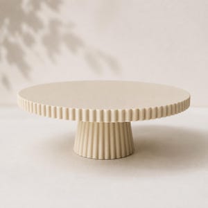 May include: A beige cake stand featuring a circular top with a fluted, scalloped edge and a matching tapered pedestal base with vertical ribbed detailing. The entire structure is finished in a solid, matte, light tan color throughout.