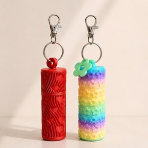 May include: Two cylindrical keychains. One is red with a heart pattern, the other is rainbow-colored with flower accents. Both have silver-colored metal clasps and rings.