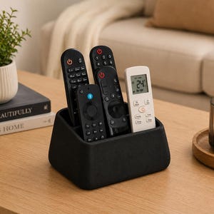 May include: A black rectangular remote control holder with four interior compartments, containing three black remote controls and one white air conditioning remote control with a digital display showing 24 degrees Celsius.