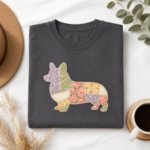 Puede incluir: Una camiseta gris oscuro que presenta un gráfico de la silueta de un perro Corgi diseñado con un patrón de patchwork. Las secciones del patchwork muestran varios estampados florales y botánicos en tonos lavanda, verde, rosa, amarillo y blanco.