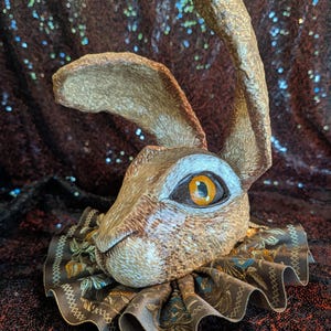 May include: A sculpted hare head with textured brown fur, large upright ears, and a prominent orange eye. The hare is adorned with a ruffled brown fabric collar featuring a repeating floral pattern in shades of blue, green, and gold.