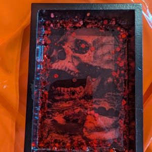 May include: A rectangular black tray featuring a central graphic of a human skull in shades of red and black. The surface is embedded with numerous small, reflective red hexagonal glitter pieces, creating a textured appearance within the frame.