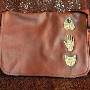 May include: A brown messenger bag featuring three distinct patches on the front flap. The top patch is a heart-shaped planchette with the text "AS ABOVE SO BELOW". The middle patch is a palmistry hand, and the bottom patch is a three-eyed cat head.