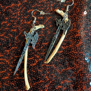 May include: A pair of dangle earrings featuring silver-toned metal axe charms suspended alongside curved, off-white bone-like pieces. The earrings are attached to standard silver-toned metal fishhook wires with clear plastic backings for secure wear.