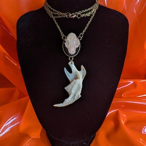 May include: A necklace featuring a bronze-toned chain, a small oval pendant with a pale, carved skull-like figure, and a large, off-white animal jawbone pendant suspended from the base of the oval piece, displayed on a black bust.