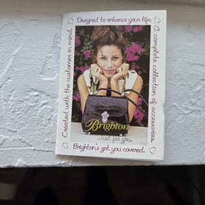 Op de afbeelding: Brochure met een vrouw en een zwarte handtas. De handtas heeft een zilveren sluiting en de merknaam "Brighton" in gouden letters. Tekst bevat "Ontworpen om je leven te verbeteren" en "Een complete collectie accessoires."