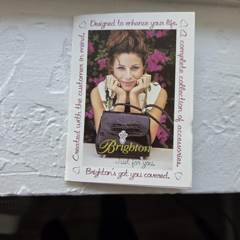 Op de afbeelding: Brochure met een vrouw en een zwarte handtas. De handtas heeft een zilveren sluiting en de merknaam "Brighton" in gouden letters. Tekst bevat "Ontworpen om je leven te verbeteren" en "Een complete collectie accessoires."