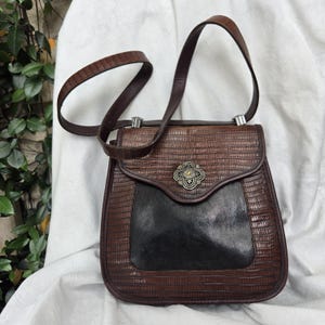 Op de afbeelding: Een vintage bruin-zwarte handtas met een textuur die op reptielen lijkt. De tas heeft een klepsluiting met een decoratieve metalen sluiting en een lange, verstelbare riem. Het onderste deel van de tas is zwart.