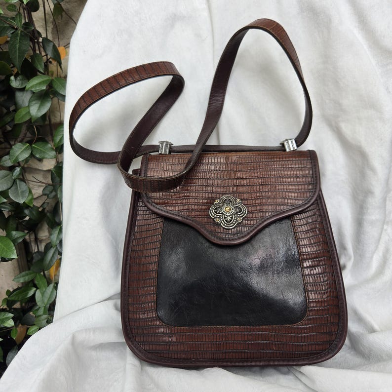 Op de afbeelding: Een vintage bruin-zwarte handtas met een textuur die op reptielen lijkt. De tas heeft een klepsluiting met een decoratieve metalen sluiting en een lange, verstelbare riem. Het onderste deel van de tas is zwart.
