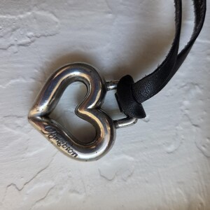 Op de afbeelding: Een zilverkleurige hartvormige hangerketting met een zwarte leren band. De hanger heeft de tekst "Brighton" gegraveerd. Het hart heeft een eenvoudig, open ontwerp.