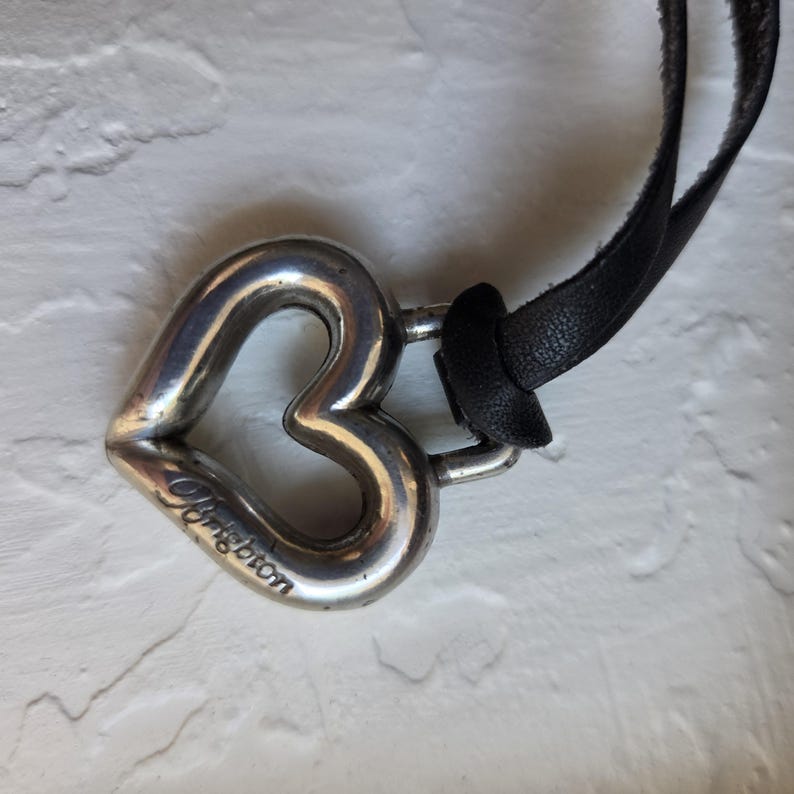 Op de afbeelding: Een zilverkleurige hartvormige hangerketting met een zwarte leren band. De hanger heeft de tekst "Brighton" gegraveerd. Het hart heeft een eenvoudig, open ontwerp.