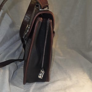 Op de afbeelding: Een donkerbruine leren handtas met een getextureerde buitenkant en een contrasterende roodbruine rand. De tas heeft een lange, verstelbare riem en een zilverkleurige sluiting. Een kleine zilveren bedel hangt aan de zijkant.