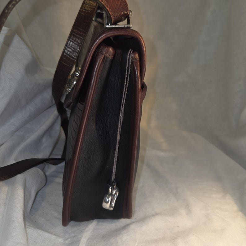 Op de afbeelding: Een donkerbruine leren handtas met een getextureerde buitenkant en een contrasterende roodbruine rand. De tas heeft een lange, verstelbare riem en een zilverkleurige sluiting. Een kleine zilveren bedel hangt aan de zijkant.