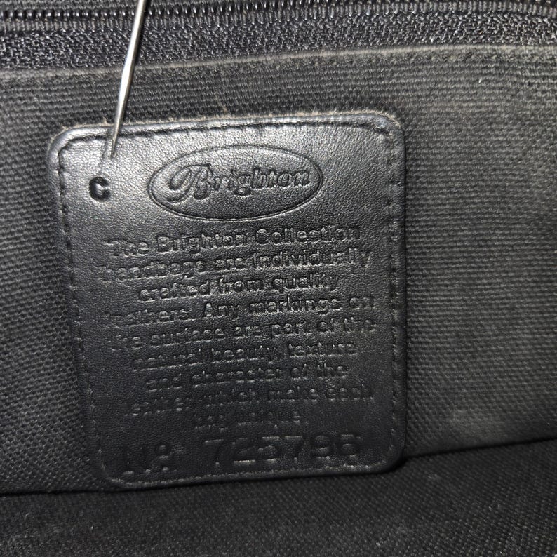 Op de afbeelding: Close-up van een zwart leren label van een Brighton handtas. Het rechthoekige label toont het Brighton-logo en de tekst: "The Brighton Collection handbags are individually crafted..." en "No. 725796".