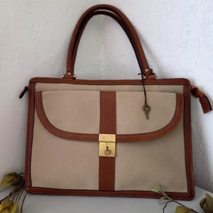 May include: A rectangular beige handbag with brown leather trim and handles. The bag features a central vertical brown leather strip with a textured pattern and a gold-toned metal clasp closure. A small gold-toned key hangs from a thin cord.