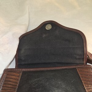 Op de afbeelding: Open zicht op een bruine en zwarte crossbodytas. De tas heeft een getextureerde bruine leren flap en afwerking, met een zwart interieur. Een zilverkleurige drukknoopsluiting is zichtbaar aan de bovenkant. Het ontwerp van de tas suggereert een vintage of klassieke stijl.