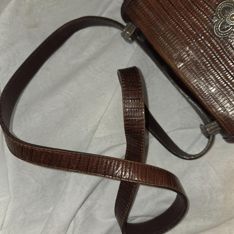 Op de afbeelding: Een bruine, getextureerde handtas met een lange, bijpassende riem. De tas heeft een decoratieve zilverkleurige sluiting en een reptielachtig patroon. De riem is gebogen en de tas lijkt van leer of een vergelijkbaar materiaal te zijn.