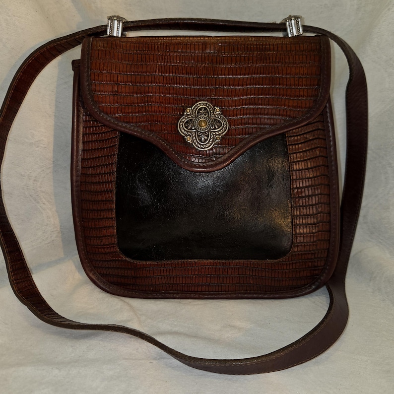 Op de afbeelding: Bruine leren crossbodytas met een getextureerd, reptielachtig patroon. De tas heeft een klepsluiting met een decoratieve zilverkleurige sluiting en een contrasterende zwarte zak. De verstelbare riem is bruin.