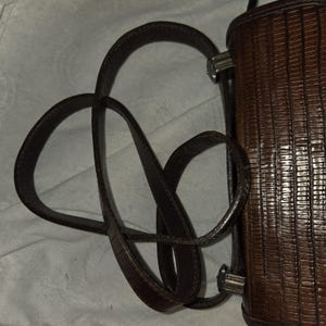 Op de afbeelding: Een bruine leren handtas met een textuur, verticaal gestreept patroon. De tas heeft een lange, lusvormige schouderriem en zilverkleurige hardware. Het ontwerp van de tas is klassiek en elegant.