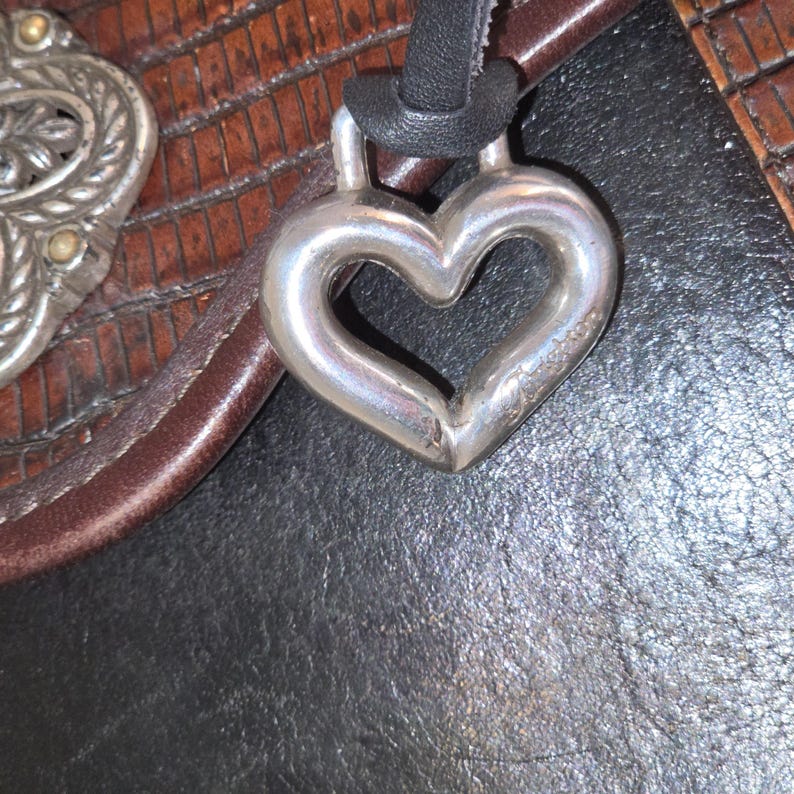 Op de afbeelding: Een zilveren hartvormige hanger hangt aan een zwart koord. De hanger is glad en glanzend, met een lichte curve. De achtergrond omvat een bruine leren riem en een zwarte, getextureerde ondergrond.