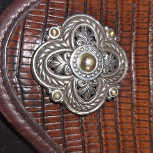 Op de afbeelding: Close-up van een decoratieve zilvermetalen versiering met een gouden midden en accenten, tegen een achtergrond van getextureerd bruin leer. De versiering heeft een bloemenontwerp met vier gebogen secties en kleine gouden studs.
