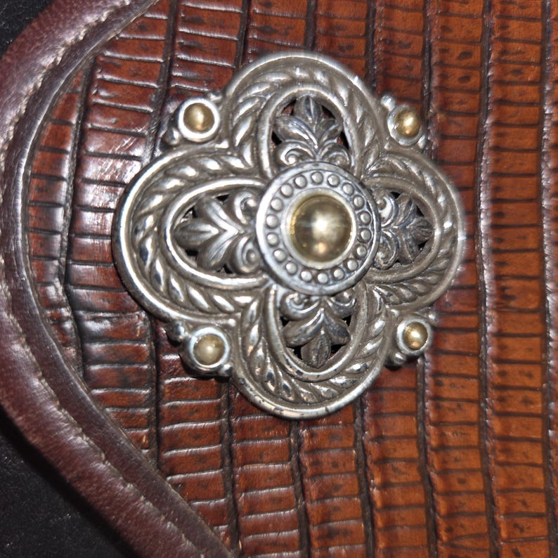 Op de afbeelding: Close-up van een decoratieve zilvermetalen versiering met een gouden midden en accenten, tegen een achtergrond van getextureerd bruin leer. De versiering heeft een bloemenontwerp met vier gebogen secties en kleine gouden studs.