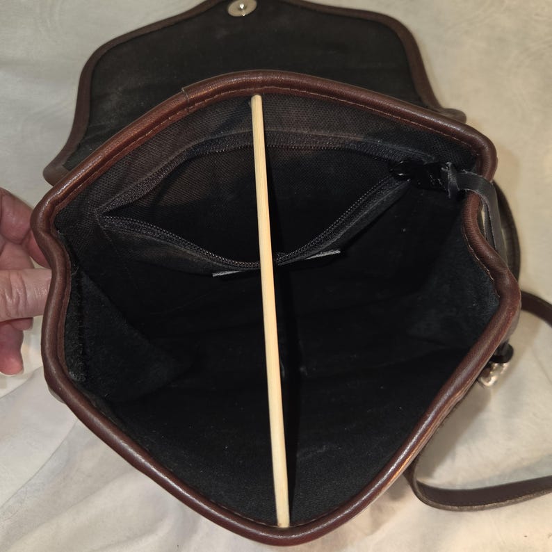 Op de afbeelding: Open zicht op een bruine leren crossbodytas met een zwart interieur. De tas heeft een klepsluiting en een binnenvak met ritssluiting. De randen zijn afgewerkt in donkerder bruin en er is een lange riem zichtbaar.