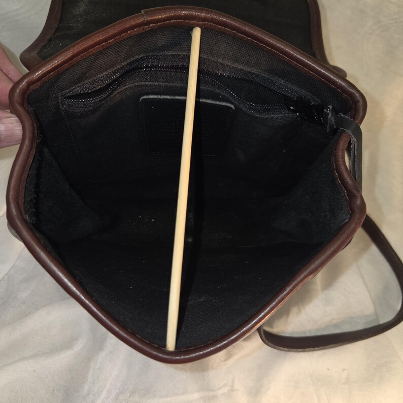 Op de afbeelding: Open zicht op een bruine leren crossbodytas met een zwart interieur. De tas heeft een klepsluiting en een lange, verstelbare riem. De binnenkant heeft een vak met ritssluiting. De randen van de tas zijn afgewerkt met donkerder bruin leer.