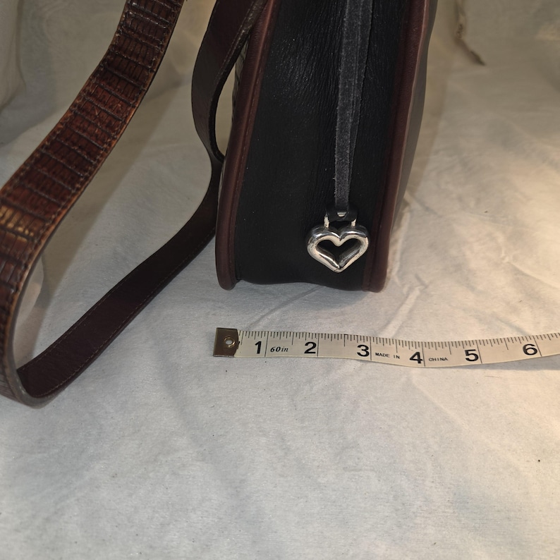 Op de afbeelding: Zwarte leren handtas met bruine rand en een bruin-beige patroonriem. Een zilveren hartvormige bedel hangt aan de zijkant. Een meetlint ligt naast de tas.