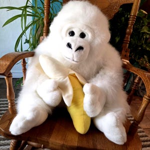 May include: A plush white gorilla stuffed animal with soft fur, black eyes, and a black nose. The gorilla is holding a bright yellow plush banana in its hands. The toy features a realistic facial structure and a seated posture with long arms.