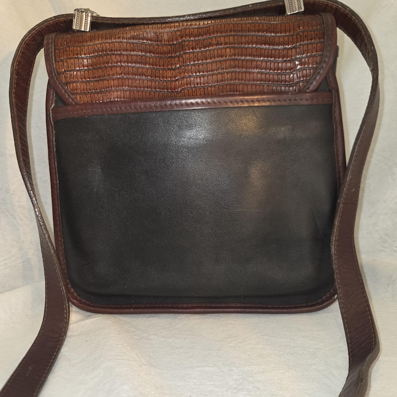 Op de afbeelding: Een bruine en zwarte leren crossbodytas met een getextureerde flap en verstelbare riem. De tas heeft een rechthoekige vorm met een contrasterende gladde zwarte leren body en een bruine, reptielachtige getextureerde flap. Zilverkleurige hardware accentueert de riem.