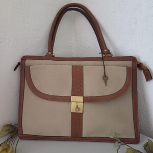 May include: A rectangular beige handbag with brown leather trim and handles. The bag features a central vertical brown textured strip, a front flap pocket, and a gold-toned metal clasp closure. A small metal key hangs from a thin cord on the handle.