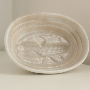 May include: Off-white ceramic serving dish with an oval shape. The interior features a raised relief design of a fish. The dish has a textured surface and a slightly cracked appearance, suggesting an aged or vintage aesthetic.