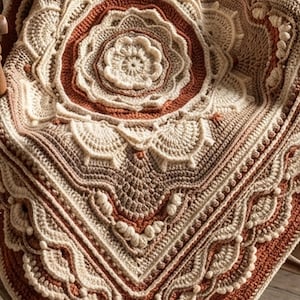 Könnte beinhalten: Eine quadratische Häkeldecke mit einem zentralen Blumenmotiv, das von komplexen geometrischen Mustern umgeben ist. Das Textil zeigt eine Farbpalette aus Creme, Beige und gebranntem Orange mit erhabenen Noppenstichen und muschelartigen Texturen.