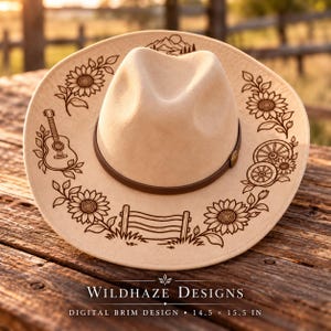 May include: A beige felt hat with a brown leather band and dark laser-engraved illustrations on the brim, including sunflowers, a guitar, a wooden fence, wagon wheels, and a mountain landscape. The text reads WILDHAZE DESIGNS 14.5 x 15.5 IN.