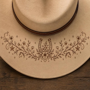 Pode incluir: Chapéu de cowboy bege com uma faixa de couro marrom. O chapéu apresenta um design floral e de ferradura marrom detalhado na aba, com estrelas, flores e cabeças de cavalo. O chapéu está sobre um fundo de madeira.