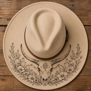 Puede incluir: Un sombrero fedora beige de ala ancha con una banda de cuero marrón oscuro alrededor de la copa. El ala está decorada con una detallada ilustración de líneas negras de un cráneo de longhorn centrado entre cactus, flores y montañas.
