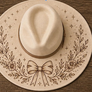 Pode incluir: Um chapéu bege de aba larga com uma faixa castanha à volta da copa. A aba está decorada com uma ilustração de linhas castanho-escuras de um laço central ladeado por dois ramos florais e estrelas de quatro pontas espalhadas.