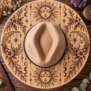 May include: A tan wide-brimmed hat featuring dark brown laser-engraved illustrations of suns with faces, crescent moons with profiles, floral motifs, and celestial phases. The hat has a structured crown and a dark brown band around the base.
