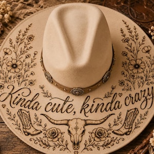 Puede incluir: Un sombrero fedora color crema con un ala decorativa con diseños florales y del oeste. El sombrero tiene una banda de cuero marrón con adornos plateados. El texto "Kinda cute, kinda crazy" es visible en el ala.