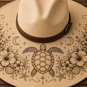 Könnte beinhalten: Beiger Fedora-Hut mit braunem Lederband. Der Hutrand zeigt ein detailliertes, geätztes Design einer Meeresschildkröte, Blumen, Sterne und Halbmonde. Ein stilvolles Accessoire.