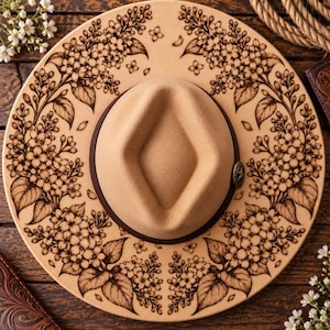 Peut inclure: Un chapeau beige à larges bords doté d'un ruban marron foncé et d'une épingle métallique en forme d'étoile. Le bord est orné d'un motif floral marron foncé détaillé représentant des grappes de lilas et des feuilles sur toute la surface.