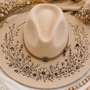 May include: A beige wide-brimmed fedora hat featuring a black ink-style floral illustration printed on the brim. The hat includes a matching beige band around the crown, secured with a small, circular metallic concho-style accent piece.