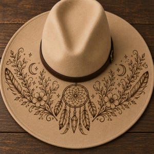 Puede incluir: Sombrero fedora beige con una banda de cuero marrón. El ala del sombrero presenta un diseño grabado detallado de un atrapasueños, plumas, flores, lunas crecientes y estrellas. El sombrero es de color neutro y tiene una forma clásica y elegante.