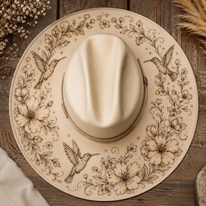 May include: Cream-colored fedora hat with a wide brim, featuring intricate floral and hummingbird designs etched around the brim. The hat has a structured crown and a thin, light-brown band.